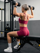Claranie One Shoulder Sports Bra in ruby red, gym scene angled back view, model seated lifting barbell, highlighting shoulder and back muscles